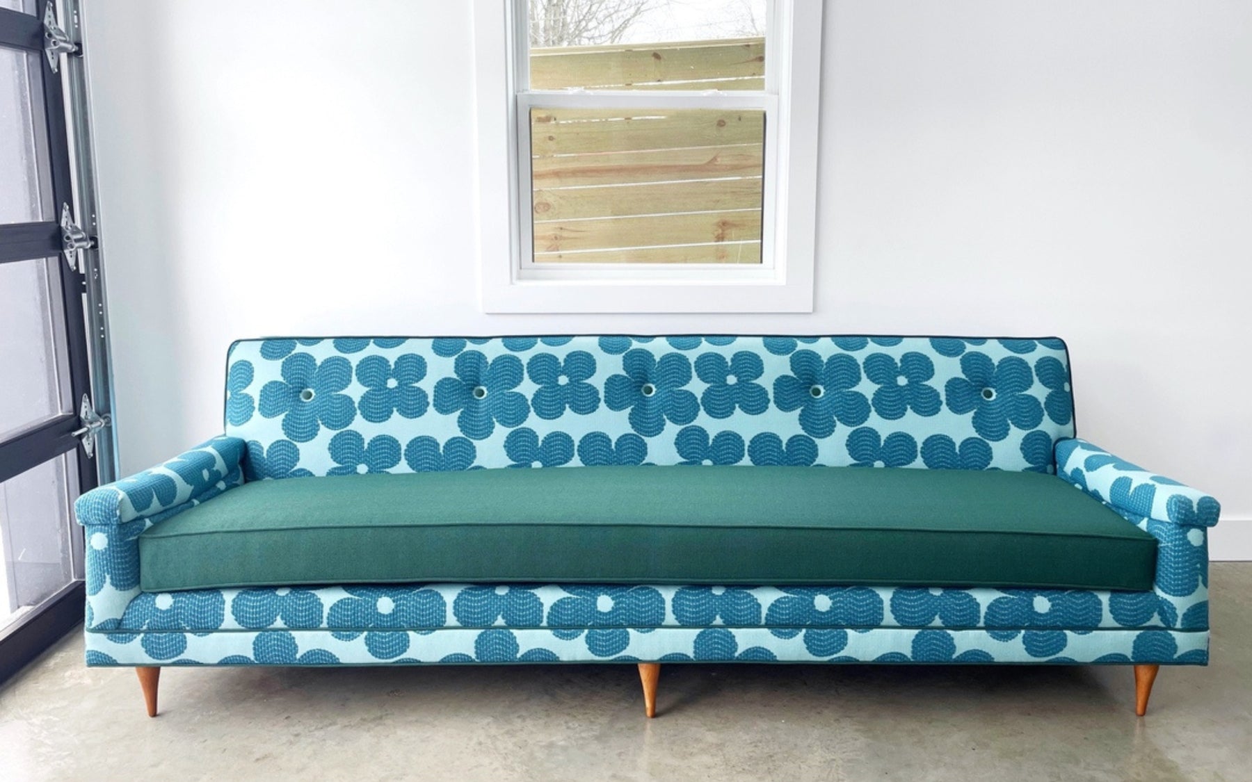 A modern-style couch reupholstered in a blue and teal floral-patterned fabric with a solid teal cushion. The couch features tapered wooden legs and is set against a white wall with a window.
