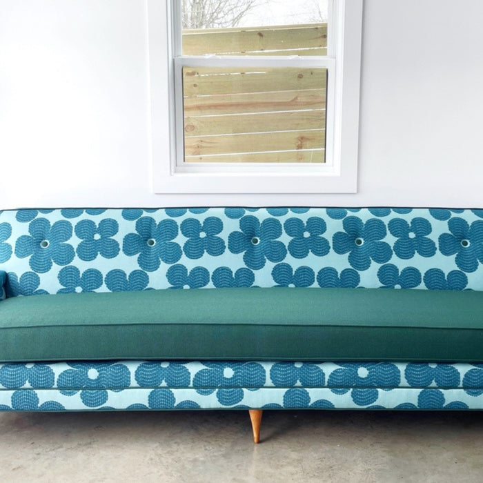 A modern-style couch reupholstered in a blue and teal floral-patterned fabric with a solid teal cushion. The couch features tapered wooden legs and is set against a white wall with a window.